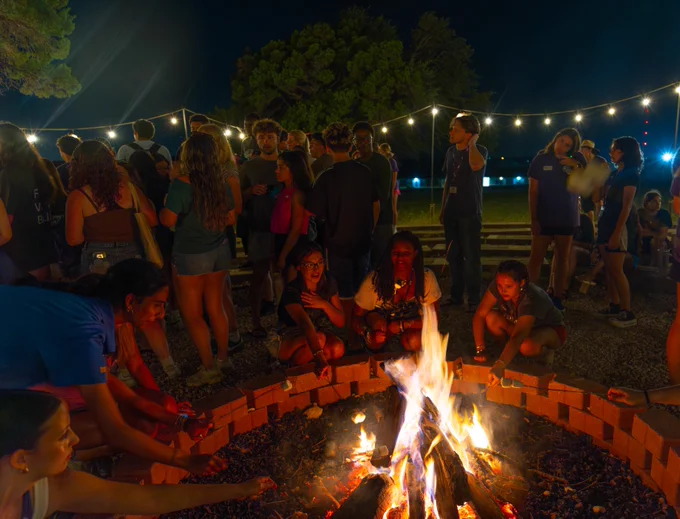 A lively group of individuals gathered around a campfire at night. String lights hang above, illuminating the scene. People are engaged in conversation, some sitting and others standing around the fire. The background is filled with trees and more participants enjoying the atmosphere.
