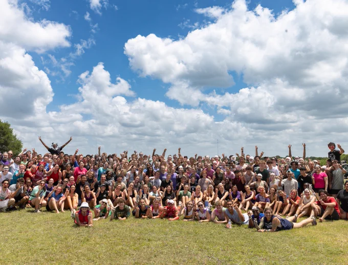 A large group of people gathered outdoors on grass, posing and cheering against a backdrop of a blue sky with scattered clouds.