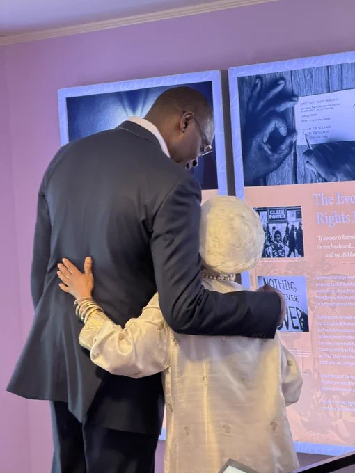 Captured from behind, the Lieutenant Governor in a dark suit has his arm warmly around the shoulder of Jawana Jackson, who is wearing a white textured headwrap and cream tunic. They are standing close together, looking at a civil rights history museum display titled "The Evolution of the Rights Movement."