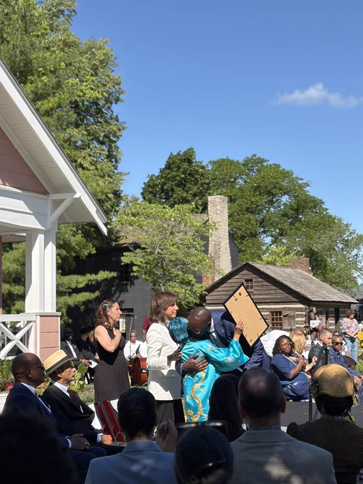 An outdoor afternoon event where the Lieutenant Governor is leaning down from a stage to warmly embrace Jawana Jackson, who is wearing a vibrant turquoise and gold patterned garment and a matching headwrap. A woman in a white blazer stands right beside them smiling, while other seated attendees and historic log cabin buildings are visible in the background under a clear blue sky.