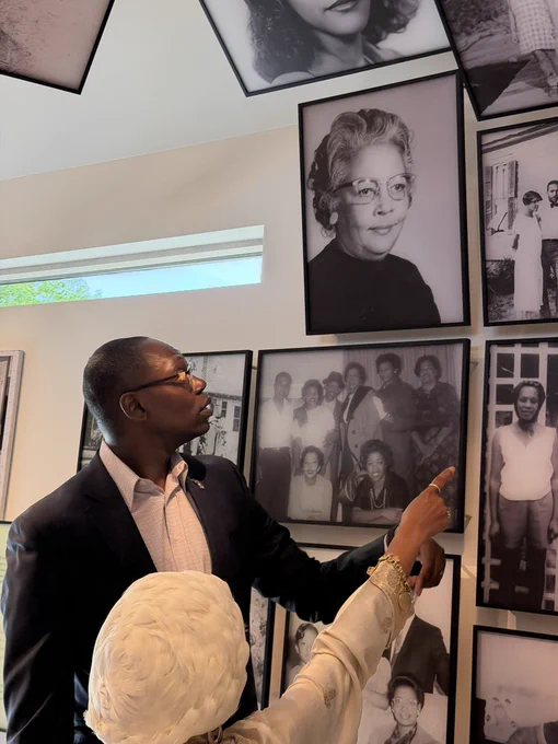 Inside a museum gallery, Jawana Jackson, seen from the back wearing her white headwrap and cream tunic, points up toward a wall of black-and-white historical portraits. The Lieutenant Governor stands beside her in his navy suit, looking up intently at the photographs she is gesturing toward.