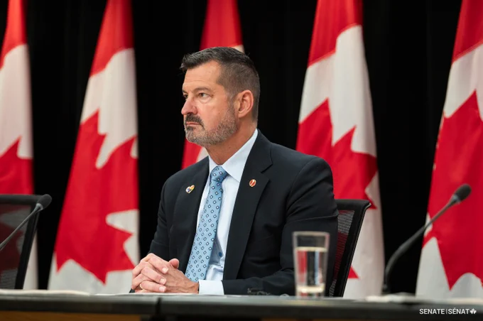 A man sitting in front of a microphone, with Canadian flags in the background.