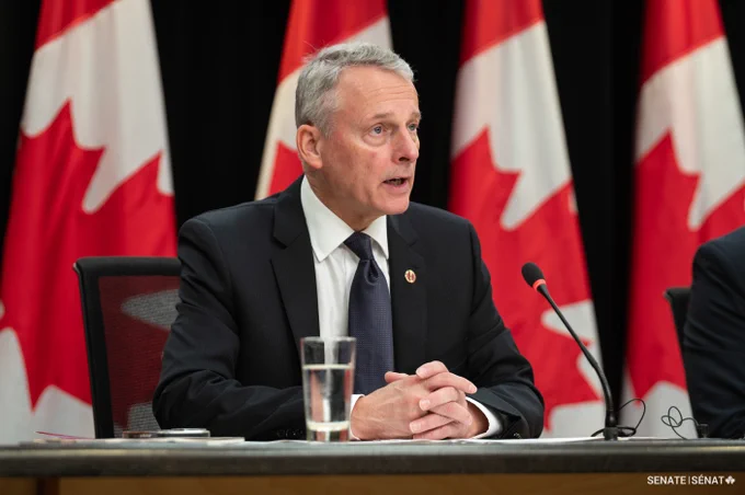 A man sitting in front of a microphone, with Canadian flags in the background.