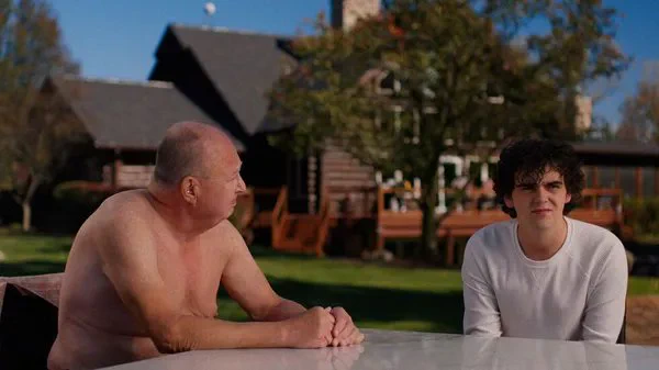 A still from the film Ponderosa. In the garden of a country house, a naked middle-aged white man sits at a table beside a slightly awkward light-skinned young man who is wearing clothes.