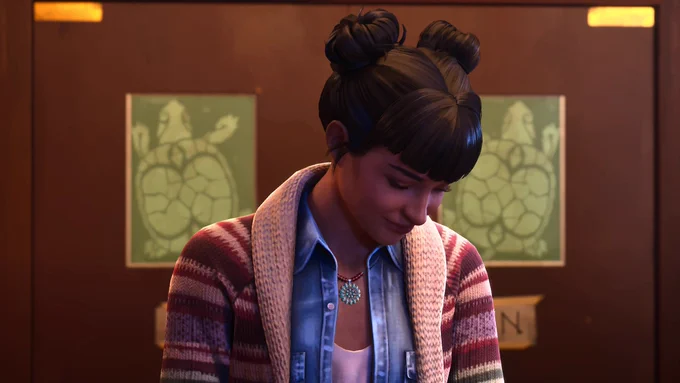 Amanda Thomas from Life is Strange: Double Exposure, an Indigenous woman of Cherokee and Algonquin descent, reacting with a shy, genuine smile after being invited on a date. Amanda is looking down bashfully, clearly happy and touched by the invitation. She is styled in her signature space buns with blunt bangs, wearing a blue denim shirt under a striped knit cardigan, and a turquoise pendant necklace. She is standing inside her bar, The Snapping Turtle, located in Lakeport, Vermont. In the background, wooden kitchen swinging doors featuring green signs with the bar's turtle logo are visible symmetrically.
