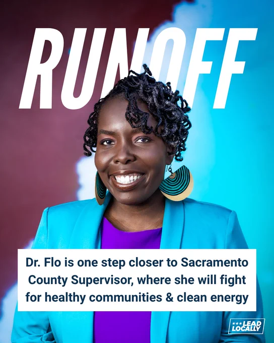 A woman with dark brown skin and black hair is smiling in a bright turquoise jacket and purple shirt.