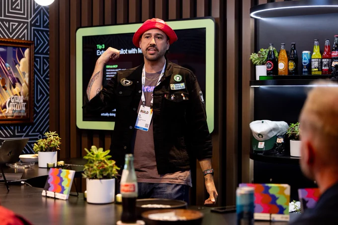 Pj behind a bar wearing a red bucket hat and a black denim jacket. He is in the middle of explaining something to folks sitting around the bar. 