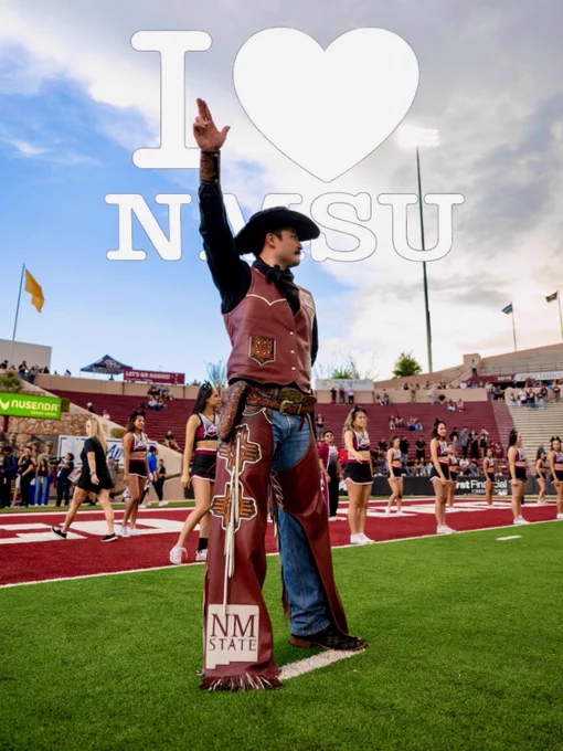 Pistol Pete stands on a football field, raising a hand. They wear a long vest with the New Mexico State University logo. In the background, cheerleaders are lined up, and a crowd is visible in the stands. Large text in the sky reads "I ♥ NMSU." 