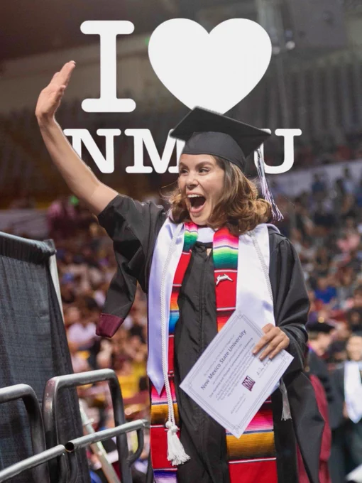 An individual in graduation attire, including a cap and gown, is joyfully waving while holding a diploma cover. They are standing in front of a large backdrop displaying the text "I ♥️ NMU" at a graduation ceremony. The venue is filled with people.