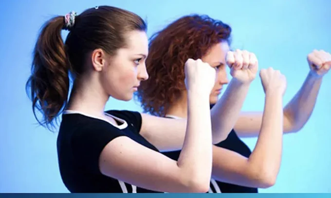 Two people practicing boxing stances side by side. The background is a soft blue, and both are wearing similar black and white tops.
