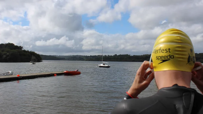 A person wearing a yellow swim cap labeled "Peterfest Speedo" stands by a lake, with cloudy skies overhead. A few small boats float on the water near a wooden dock.