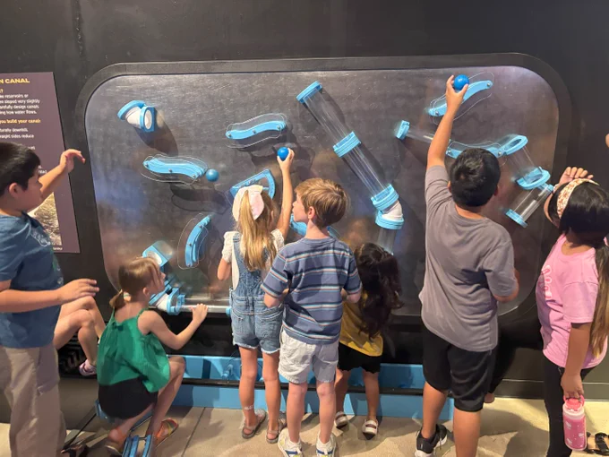 Children interact with a large wall-mounted maze of transparent tubes and blue connectors, exploring how pieces fit together.