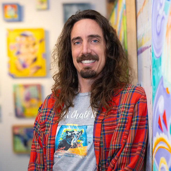 A smiling man with long hair stands in an art gallery, wearing a red plaid shirt over a graphic tee with lively artwork in the background. The mood is vibrant and creative.