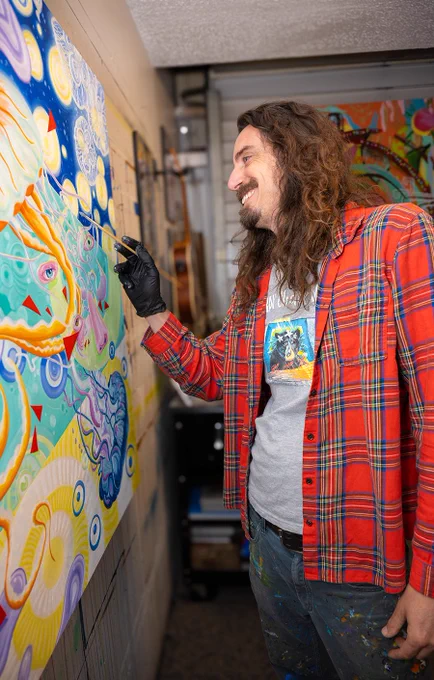 A focused artist works on a vibrant abstract painting, smiling in a studio. The lively colors and intricate designs reflect a creative, joyful mood.