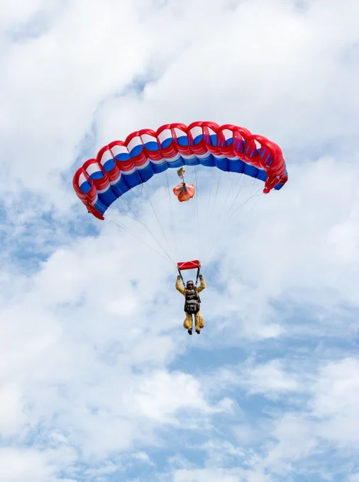 Smokejumper descending with a red, white, and blue parachute against a partly cloudy sky.