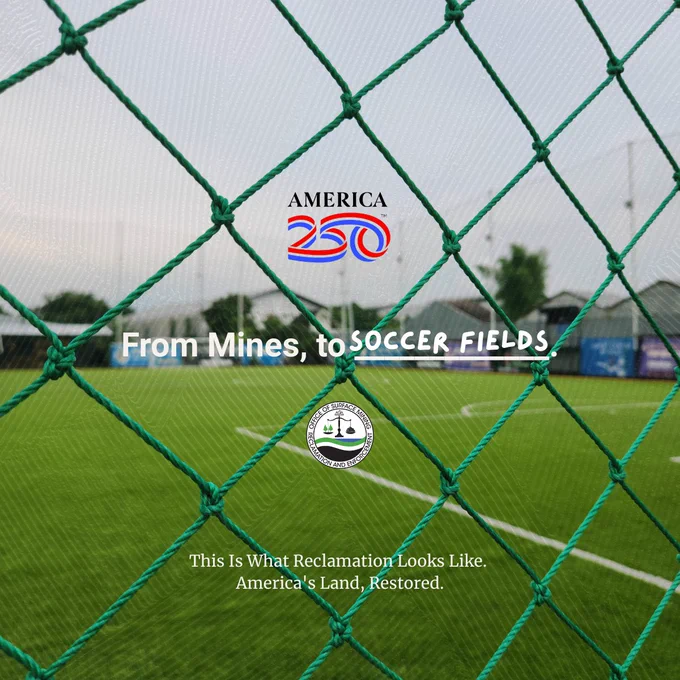 Green netting in focus with a blurred soccer field and goalposts in the background under a cloudy sky.