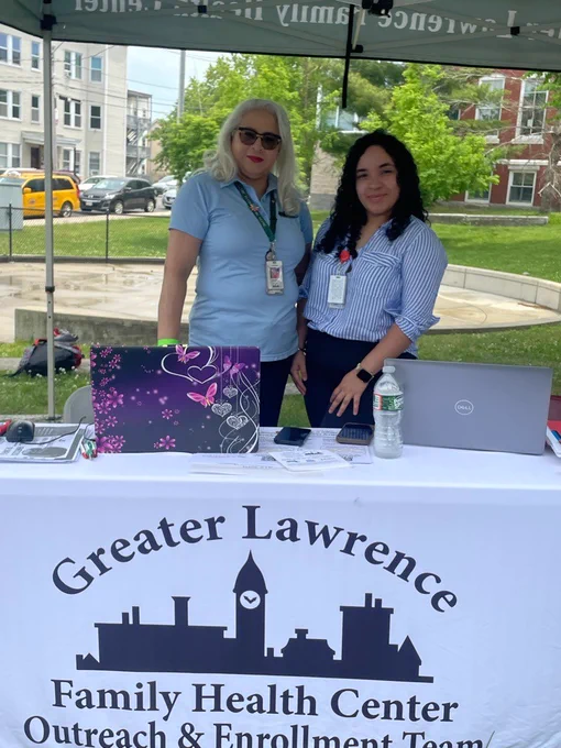 Two GLFHC staff standing behind a table with laptops and water bottle at Greater Lawrence Family Health Center outreach event outdoors.
