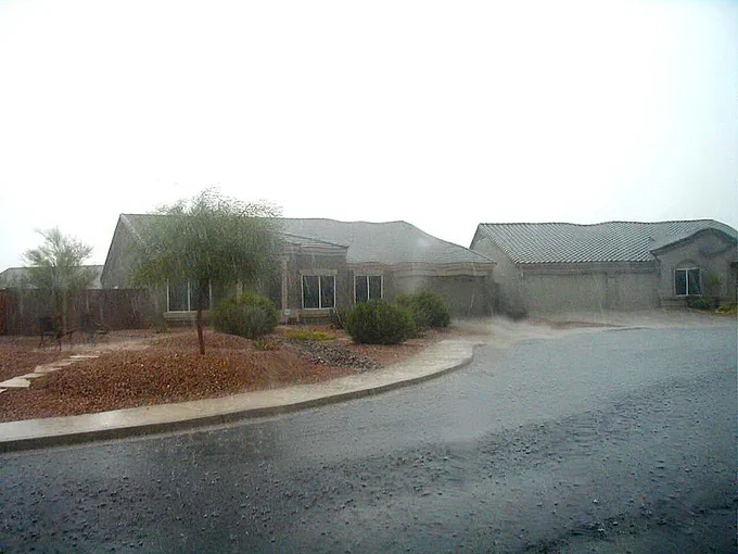 Single-story suburban house with a tiled roof and desert landscaping under heavy rain.