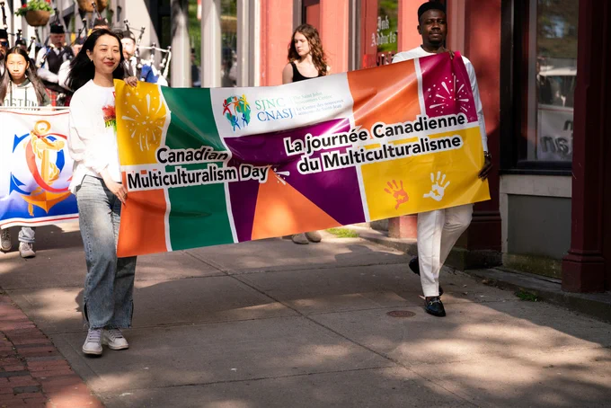 Two people holding a colorful banner promoting Canadian Multiculturalism Day during a street parade or event.