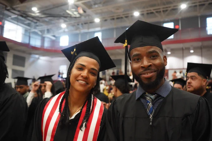 Two graduates wearing caps and gowns stand together inside a gymnasium filled with other graduates and attendees.