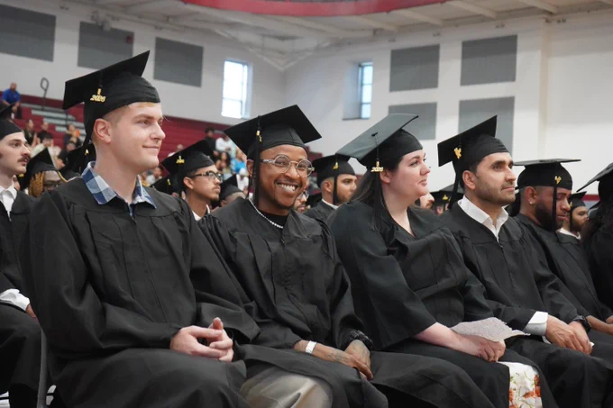 A group of graduates wearing black caps and gowns seated in a gymnasium during a commencement ceremony.