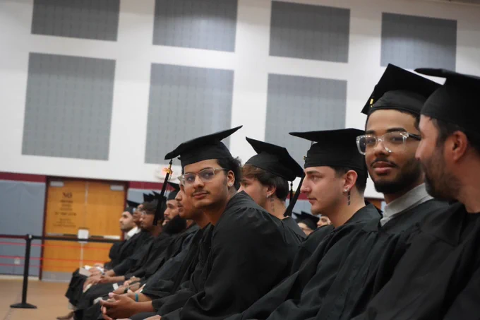 Row of graduates in black caps and gowns seated indoors during a ceremony.