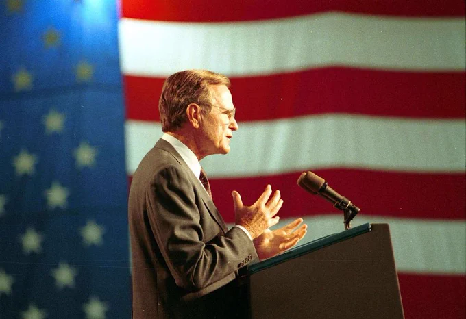 Former President George H.W. Bush speaking at a podium in front of a large United States flag.