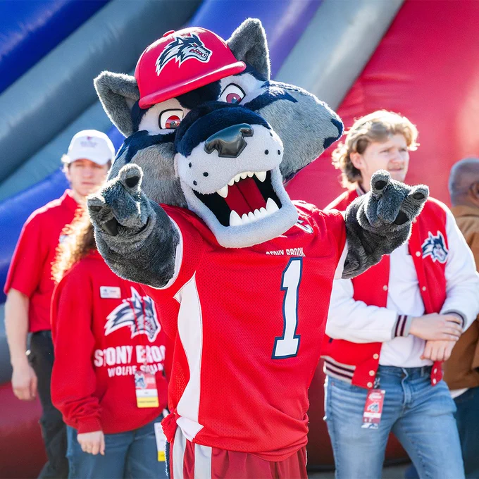 Wolfie points toward the camera during a Stony Brook event while standing in front of an inflatable attraction.