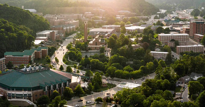 A scenic aerial view of the Appalachian State University campus in Boone, North Carolina. The image shows numerous university buildings surrounded by lush greenery, with roads winding through the campus. Sunlight filters through, casting a warm glow over the landscape.
