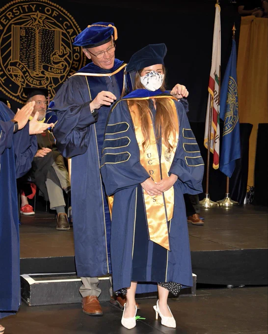 Heather being hooded during the PhD graduation ceremony. She is wearing an N95 respirator with blue flowers to match her University of California gown