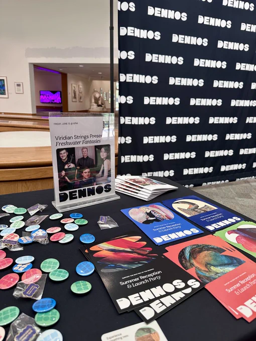 A table is set up with promotional materials and buttons. A sign displays an event called "Viridian Strings Present Freshwater Fantasies" at the Dennos Museum Center. The backdrop features the word "Dennos" repeated. Colorful brochures are scattered across the table.