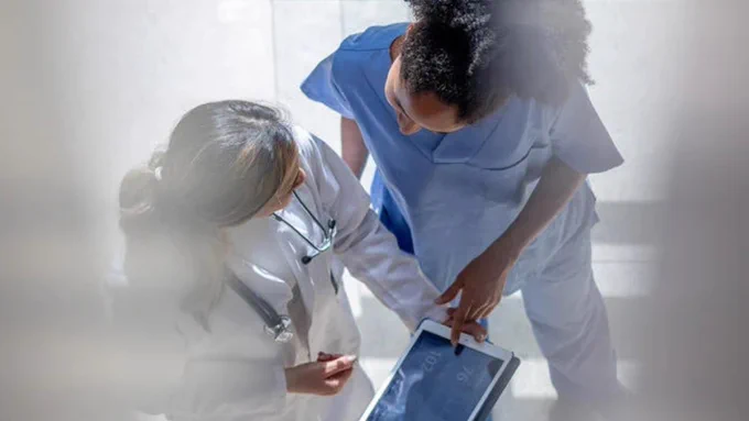 Doctor talking to a nurse in the corridor of a hospital while pointing at a digital tablet.