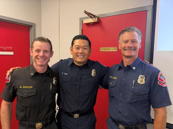 Newly promoted Captain Keita Sato poses for a photo with Battalion Chief Bryan Kistler and Captain Berkley Northrop during a Huntington Beach Fire Department promotion ceremony. The three department leaders stand together smiling inside a department meeting room.