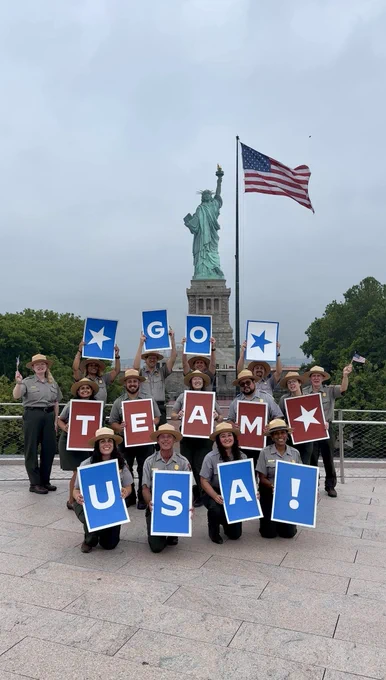 A group of National Park Service employees stands on a rooftop with the Statue of Liberty and a large American flag in the background. The employees hold red, white, and blue signs that spell “TEAM USA!” while smiling and waving toward the camera under an overcast sky.