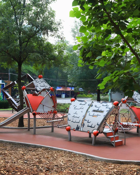 Parque infantil con estructuras metálicas geométricas en rojo y gris, rodeadas de árboles y madera triturada en el suelo.