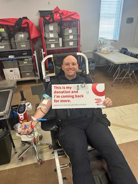 A uniformed police officer sits in a reclining chair while donating blood. The officer is holding a sign that reads: “This is my 1st donation and I’m coming back for more,” and “Experience what you get when you give."