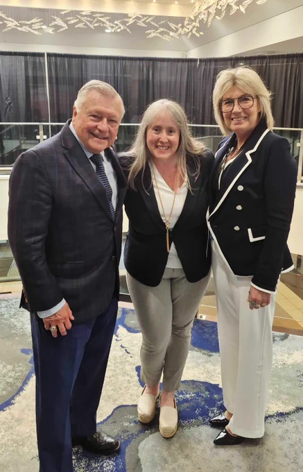 A man in a dark plaid suit and two women, one in a black blazer and grey pants and the other in a black and white blazer and white pants, posing together at an event.
