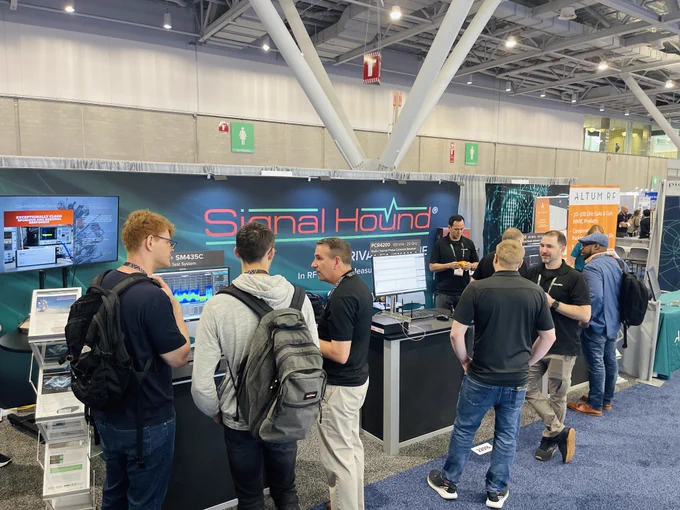 A group of people are gathered at a trade show booth for Signal Hound. The booth features electronic equipment and display screens with graphs and data. There are several participants interacting, some with backpacks. The setting is a spacious indoor trade show venue with high ceilings and large lighting fixtures.
