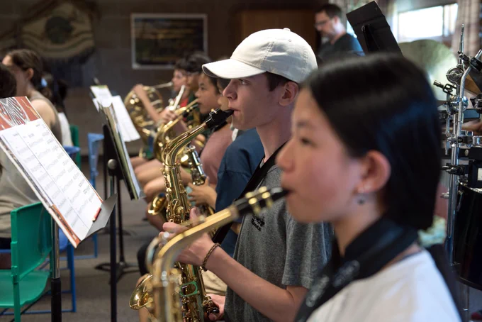 Group of young musicians seated indoors in a rehearsal space, playing saxophones and brass instruments while reading sheet music on stands.