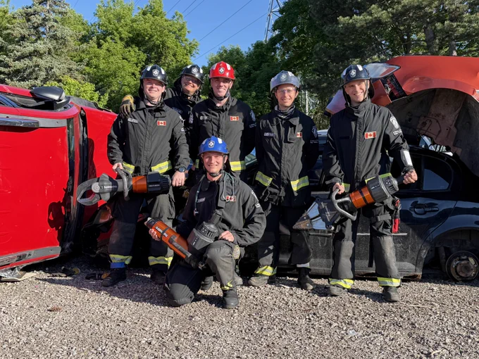 A group of six Burlington firefighters in protective gear are posing outdoors with hydraulic rescue tools. They are standing in front of two damaged vehicles, a red one overturned and a black one damaged. There are green trees and a clear sky in the background.