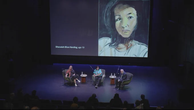 A photo of three people sitting on a stage in front of an audience, while a photo of a portrait is projected behind them.