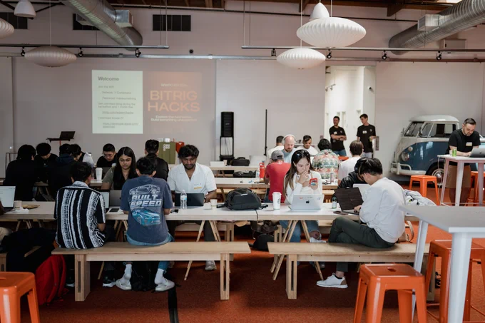 A bustling tech event scene with participants focused on laptops at communal tables, a presentation on Bitrig Hacks in the background.