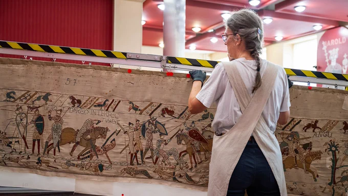 A person in an apron and gloves carefully moves a large tapestry in a museum