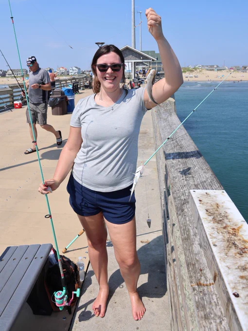 A person on a pier holding a fishing rod and displaying two small fish. The pier overlooks a body of water, with a beach and additional structures visible in the background. The scene is sunny, and the person is smiling.