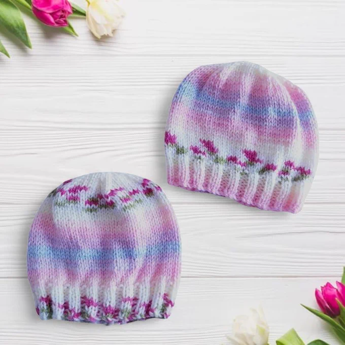 Two knitted beanies lie side by side on pale whitewashed wood. They feature a soft variegated blend of pink, purple, blue and white yarn, with delicate floral-style patterns in darker pink and green near the ribbed cuffs. Fresh tulips in pink, white and yellow with green leaves frame the corners, creating a bright, delicate and fresh scene.