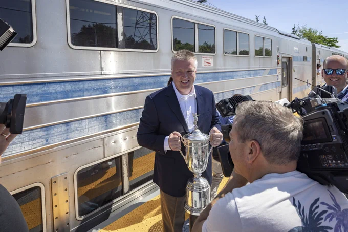 LIRR President Rob Free presents the trophy with a train in the background and cameramen in the foreground.