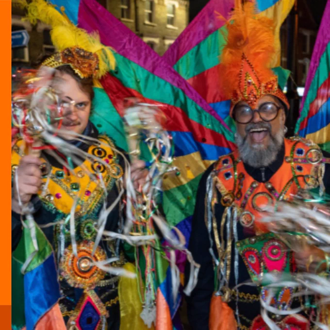 Two men in vibrant, sequined carnival costumes with feathered headdresses, celebrating at night.