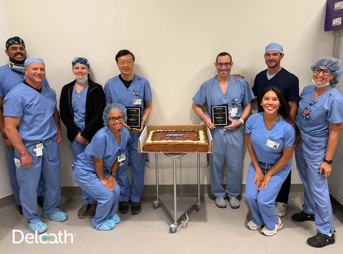 Clinical team members in surgical scrubs gather around a cake and recognition plaques to celebrate Delcath's 100th treatment case milestone at a hospital treatment center.