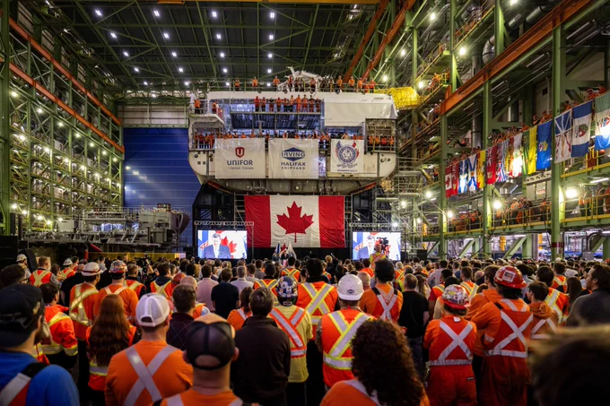 Large crowd of workers in orange safety vests gathered inside a shipyard for a ceremony with a giant Canadian flag displayed on a ship's hull.