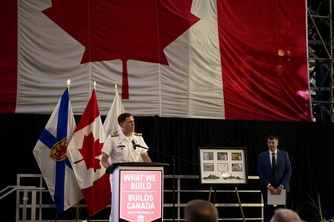 Canadian naval officer speaking at a podium with a large Canadian flag and provincial flags behind him during a formal event.